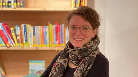 Eine Lehrerin hält lächelnd ein Buch vor einem Bücherregal