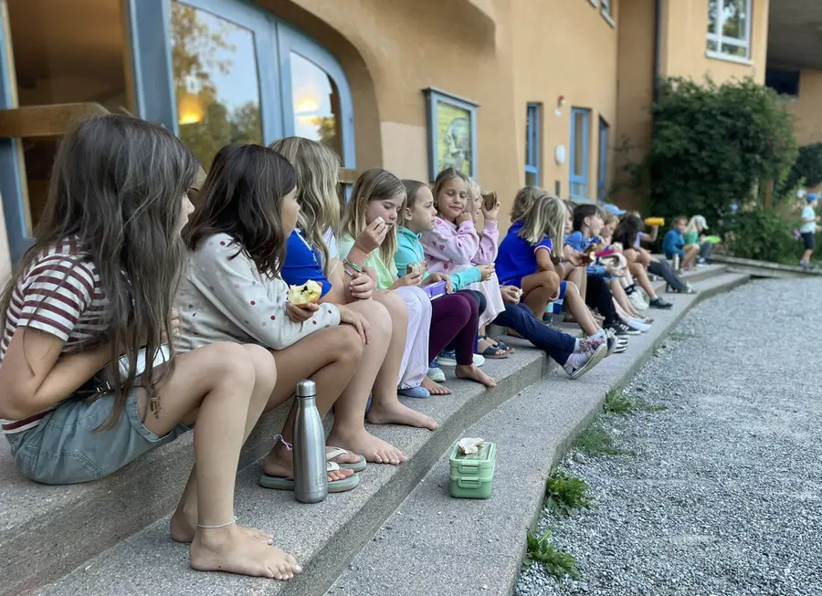 Schülerinnen und Schüler sitzen barfuß auf den Stufen vor dem Schulgebäude und essen ihr Frühstück