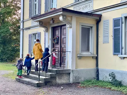Kinder gehen mit einer Erwachsenen die Treppe zur Eingangstür der Kindergarten-Villa hinauf