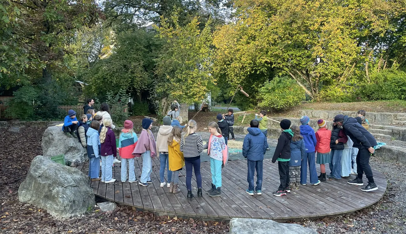 Eine Schulklasse steht im Kreis auf der runden Holzbühne des Schulhofs