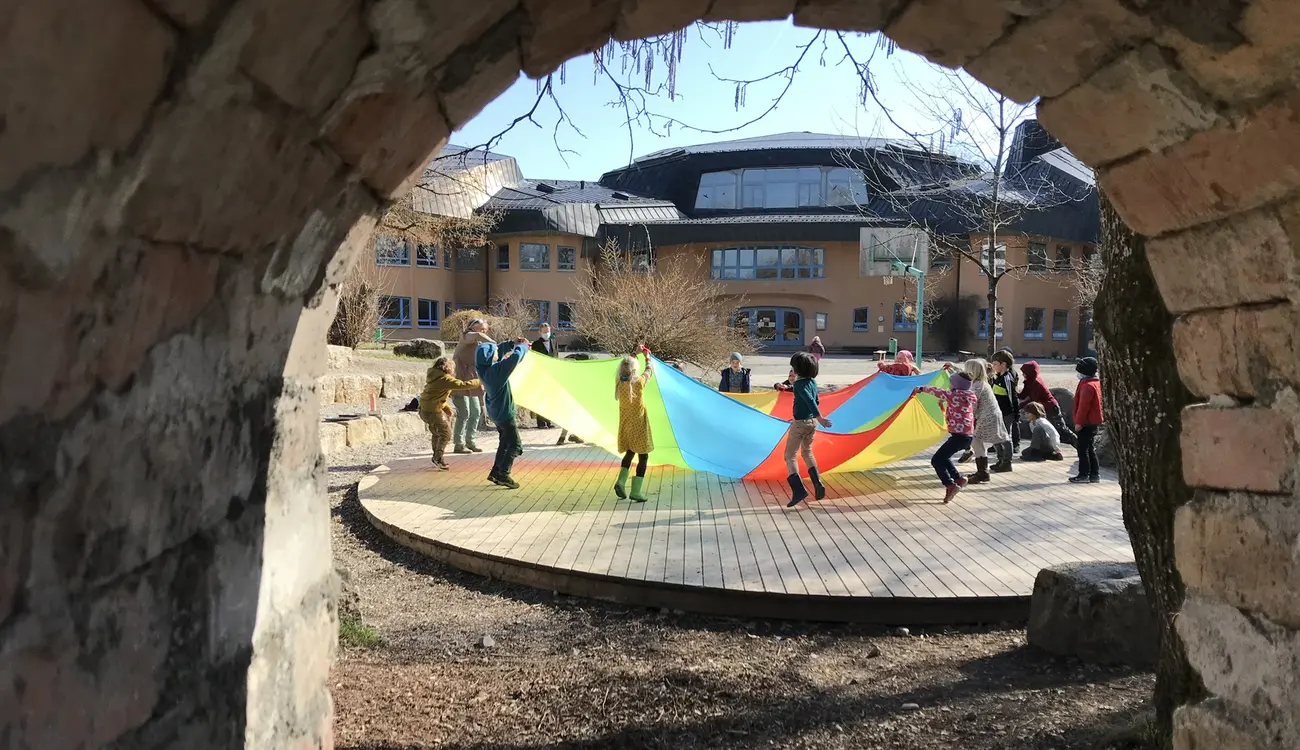 Kinder spielen mit einem bunten Schwungtuch auf dem Schulhof, durch einen Steinbogen gesehen
