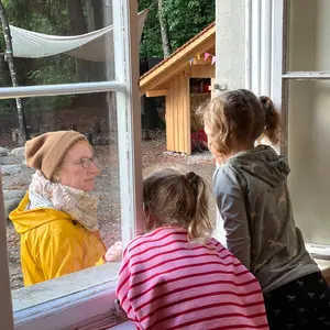 Zwei Kinder schauen aus dem offenen Fenster zu einer Frau in gelber Regenjacke im Garten
