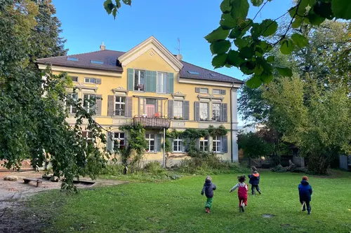 Kinder laufen über die Wiese vor der gelben Villa des Hauskindergartens