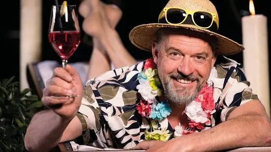 Ein Mann mit Strohhut, Hawaiihemd und Blumenkette prostet mit einem Glas zu